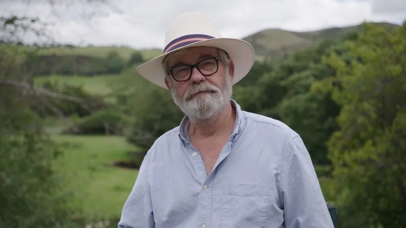 A man in a sunhat and glasses, with green hills behind him